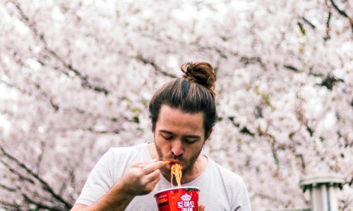 Man standing around cherry blossom trees, eating ramen.